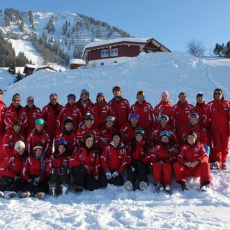 A group of skiers in red suits is posing in the snow. In the background, snow-covered mountains and cabins can be seen. | © Skischule Riezlern GmbH | Frank Felder