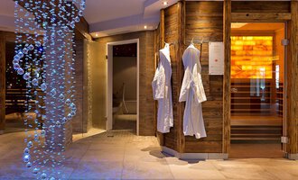 Ein modernes Wellness-Interior mit sanfter Beleuchtung. Links hängen weiße Bademäntel, und eine beeindruckende Wasserinstallation verleiht dem Raum ein entspannendes Ambiente. | © Aparthotel Kleinwalsertal Haller GmbH & Co KG