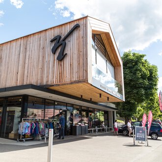 Ein modernes Geschäft mit Holzfassade und großen Fenstern. Im Vordergrund sind Kleidung und Fahrräder ausgestellt, umgeben von einer schönen Berglandschaft. | © Sport Kessler GmbH