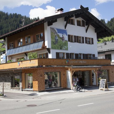 Ein charmantes Gebäude mit traditioneller Architektur, umgeben von Bergen. Im Erdgeschoss befindet sich ein Geschäft mit großen Fenstern. | © Sport Pauli Matthias Bischof e.U
