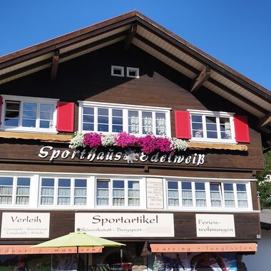 Ein traditionelles Holzhaus mit roten Fensterläden und Blumenschmuck. Der klare Himmel und die umliegenden Berge sorgen für eine idyllische Atmosphäre. | © Sport Edelweiß | N. Lughammer