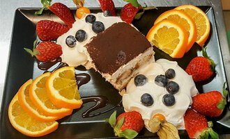 Ein schönes Dessert auf einem schwarzen Teller. Es besteht aus Tiramisu, Sahne und frischen Früchten wie Erdbeeren und Orangen.