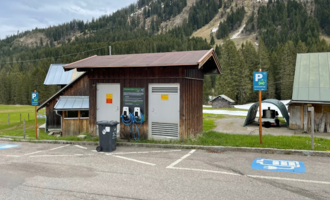 A small wooden building with charging stations for electric cars is located in an outdoor parking lot. In the background, green meadows and wooded hills can be seen. | © Kleinwalsertal Tourismus