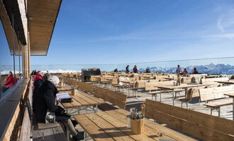 Ein idyllisches Restaurant auf einer Bergterrasse mit einer Vielzahl von Holztischen und Stühlen. Im Hintergrund sind schneebedeckte Berge und ein klarer Himmel zu sehen. | © OBERSTDORF · KLEINWALSERTAL BERGBAHNEN