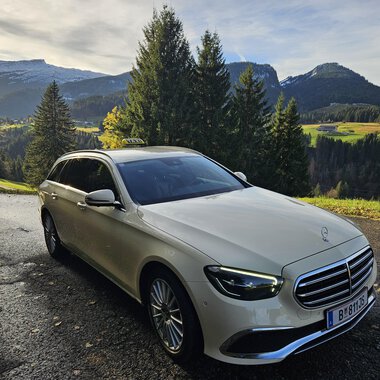 An elegant car is standing on a picturesque road in the mountains. In the background, green meadows and wooded hills can be seen. | © Taxi Beranek | Elmar Beranek