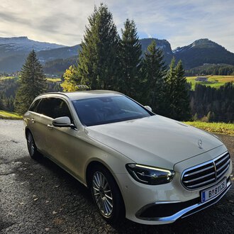 An elegant car is standing on a picturesque road in the mountains. In the background, green meadows and wooded hills can be seen. | © Taxi Beranek | Elmar Beranek