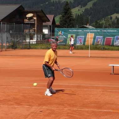 A man is playing tennis on a red court with a view of the mountains. In the background, other players and a tennis wall can be seen. | © Sportverein Kleinwalsertal