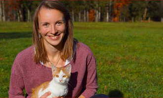 A smiling woman is sitting on a meadow holding an orange-and-white cat. Autumn trees can be seen in the background. | © Tierarztpraxis Oberstdorf | Regina Baumgartner