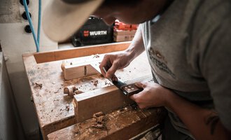Ein Handwerker arbeitet mit einem Werkzeug an einem Holzstück. Späne liegen auf der Werkbank, während er präzise Schnitte macht. | © Türtscher Die Schreinermanufaktur