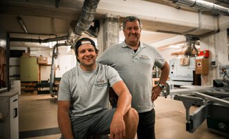 Two men are standing in a workshop. The younger man is sitting on a stool, while the older man stands proudly next to him. | © Türtscher Die Schreinermanufaktur