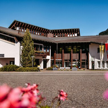 Ein modernes Gebäude mit einer eleganten Fassade und umgeben von schönen Blumen. Im Hintergrund sind Berge und ein klarer Himmel sichtbar. | © Walser Raiffeisen Bank AG