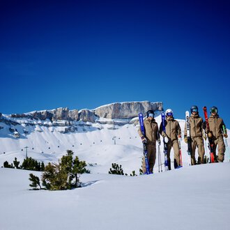 A group of skiers stands on a snow-covered area. In the background, you can see impressive mountain peaks and a clear blue sky. | © Skischule Jörihalde Kleinwalsertal | Herbert Jochum