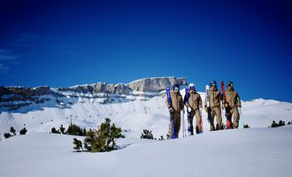 A group of skiers stands on a snow-covered area. In the background, you can see impressive mountain peaks and a clear blue sky. | © Skischule Jörihalde Kleinwalsertal | Herbert Jochum