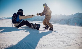 Two snowboarders on a snowy slope. One is helping the other, who is sitting on the ground. | © Skischule Jörihalde Kleinwalsertal | Ole Zumpolle