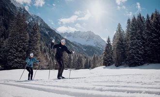 Two people cross-country skiing in a snowy landscape. In the background, majestic mountains and a clear, sunny sky can be seen. | © Skischule Jörihalde Kleinwalsertal | Ole Zumpolle