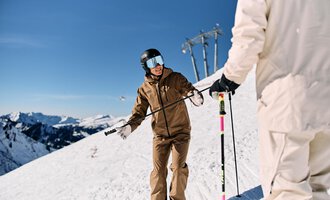 A skier in a brown ski jacket is standing on a snow-covered slope. In the background, there are snow-covered mountains and a blue sky. | © Skischule Jörihalde Kleinwalsertal | Oliver Farys