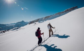 Two hikers are walking through deep snow in the mountains. The sun is shining clearly in the blue sky. | © Skischule Jörihalde Kleinwalsertal | Ole Zumpolle