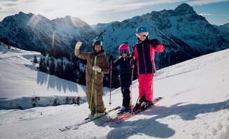Three skiers are standing on a snowy slope and waving at the camera. In the background, impressive mountains and a clear sky can be seen. | © Skischule Jörihalde Kleinwalsertal | Ole Zumpolle
