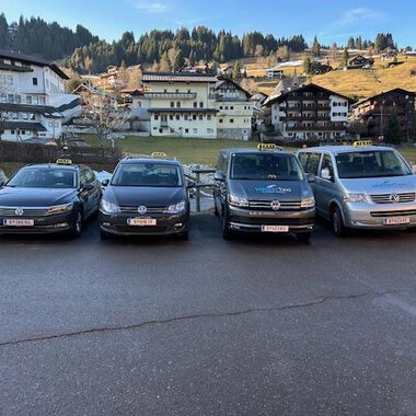 Four taxis are lined up in front of a picturesque mountain landscape. In the background, traditional alpine buildings and wooded hills can be seen. | © Walser Taxi | Josef Düringer