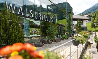 Das Walserhaus ist ein modernes Gebäude mit großen Fenstern in einer malerischen Umgebung. Im Vordergrund sind bunte Blumen und ein Wanderer zu sehen. | © Kleinwalsertal Tourismus | Oliver Farys