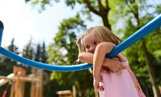 Ein kleines Mädchen spielt auf einem Spielplatz. Sie hält sich an einem blauen Spielgerät fest und schaut fröhlich. | © Kleinwalsertal Tourismus | Oliver Farys
