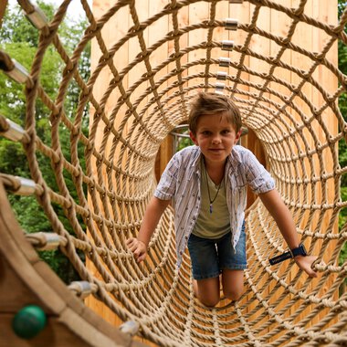 A boy is crawling through a large net on a playground. Trees can be seen in the background. | © Kleinwalsertal Tourismus | Oliver Farys