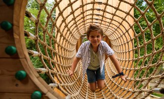 Ein Junge krabbelt durch ein großes Netz auf einem Spielplatz. Im Hintergrund sind Bäume zu sehen. | © Kleinwalsertal Tourismus | Oliver Farys