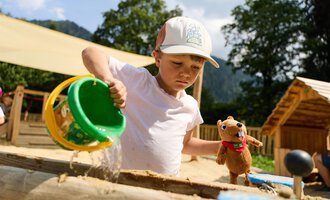 Ein Junge im Sandkasten gießt Sand aus einem Eimer. Neben ihm hält er ein Plüschspielzeug. | © Kleinwalsertal Tourismus | Oliver Farys