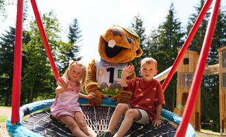 Ein fröhlicher Spielplatz mit zwei Kindern und einem Maskottchen. Sie sitzen in einer großen Schaufelwippe und genießen die Sonne. | © Kleinwalsertal Tourismus | Oliver Farys