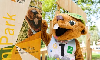 Ein lustiger Maskottchen-Biber steht neben einem Schild des BurmiParks. Der Biber trägt ein grünes Hüte und ein sportliches Outfit. | © Kleinwalsertal Tourismus | Oliver Farys