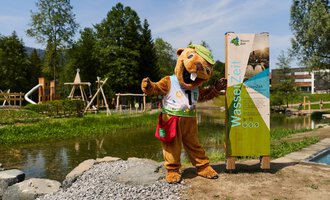 Ein freundliches Maskottchen in Tierkostüm steht neben einem Informationsschild am Wasser. Im Hintergrund sind Spielgeräte und viel Grün zu sehen. | © Kleinwalsertal Tourismus | Oliver Farys