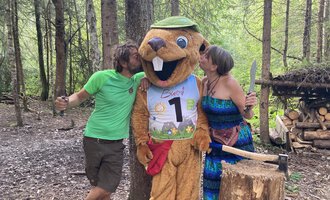 A funny mascot in the shape of a beaver stands between two people in the forest. The humans seem to be having fun and are kissing the mascot on the cheeks. | © Wildnisschule Kleinwalsertal