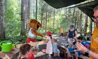 A cute mascot greets children in the forest. Some adults and children are sitting at a table and enjoying their time in nature. | © Wildnisschule Kleinwalsertal