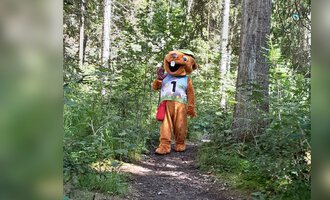 A mascot in a forest is wearing a jersey number. It stands on a narrow path between the trees. | © Wildnisschule Kleinwalsertal