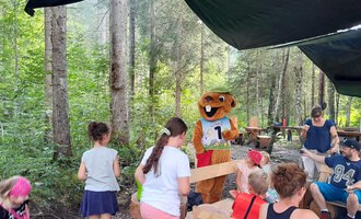 Eine Gruppe von Kindern steht in einem Wald und sieht einen Maskottchen, das einen Wettkampf über eine Zahl hält. Die fröhliche Stimmung wird durch das grüne Umfeld verstärkt. | © Wildnisschule Kleinwalsertal
