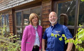 Ein Mann und eine Frau stehen lächelnd vor einem Holzgebäude. Die Frau trägt eine pinke Jacke und der Mann ein blaues Hemd. | © Wirtshaus Hoheneck | Klaus Ruland
