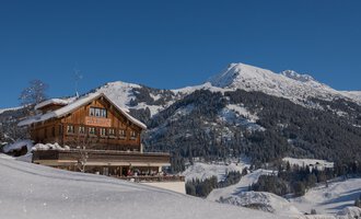 Ein rustikales Chalet im Schnee, umgeben von majestätischen Bergen. Der klare blaue Himmel lässt die Winterlandschaft besonders strahlen. | © Wirtshaus Hoheneck | Klaus Ruland