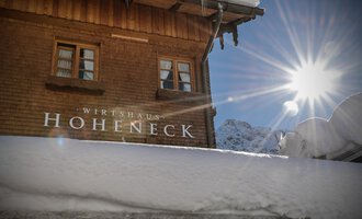 Ein gemütliches Holzhaus mit der Aufschrift „Wirtshaus Hoheneck“ unter einer Schneedecke. Die Sonne strahlt hell über die verschneite Umgebung.