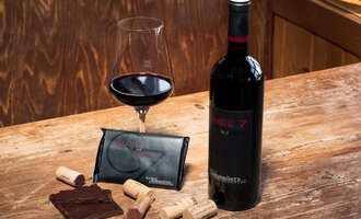 Eine Flasche Rotwein mit einem Weinglas, Schokolade und Korken auf einem Holztisch. Der Wein ist in einer eleganten Verpackung präsentiert. | © Wirtshaus Hoheneck | Klaus Ruland