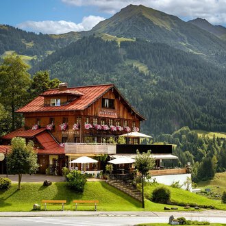 A rustic chalet in the mountains with a red roof and many flowers. The surroundings are green and hilly, surrounded by majestic mountains.