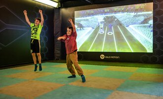 Zwei Kinder springen fröhlich in einem Spielraum. Im Hintergrund ist eine große Projektionsleinwand mit einem interaktiven Spiel sichtbar. | © Wonniland Sonthofen | interSPA Gruppe