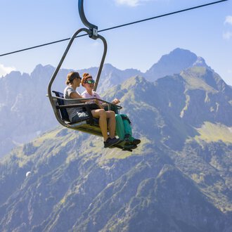 Zwei Personen sitzen im Sessellift und genießen die Aussicht auf die Berge. Im Hintergrund sind grüne Wiesen und hohe Gipfel zu sehen. | © Kleinwalsertal Tourismus | Frank Drechsel