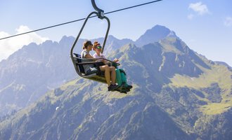 Zwei Personen sitzen im Sessellift und genießen die Aussicht auf die Berge. Im Hintergrund sind grüne Wiesen und hohe Gipfel zu sehen. | © Kleinwalsertal Tourismus | Frank Drechsel