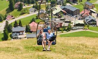 Zwei Frauen sitzen in einer Sesselliftbahn und fahren eine Bergwiese hinauf. Im Hintergrund sind grüne Hügel und ein kleines Dorf mit Häusern zu sehen. | © Kleinwalsertal Tourismus | Oliver Farys