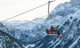 Eine Person fährt in einer Sesselbahn über schneebedeckte Berge. Die Landschaft ist majestätisch und zeigt die winterliche Schönheit der Alpen. | © Kleinwalsertal Tourismus | Oliver Farys
