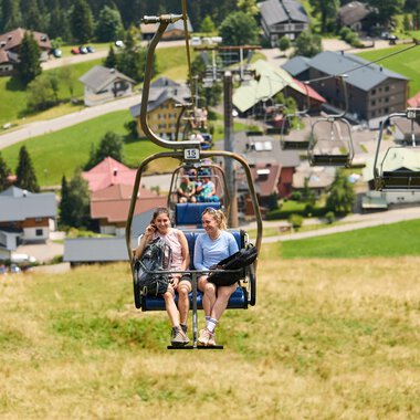 Zwei Frauen sitzen in einer Sesselliftbahn und fahren eine Bergwiese hinauf. Im Hintergrund sind grüne Hügel und ein kleines Dorf mit Häusern zu sehen. | © Kleinwalsertal Tourismus | Oliver Farys