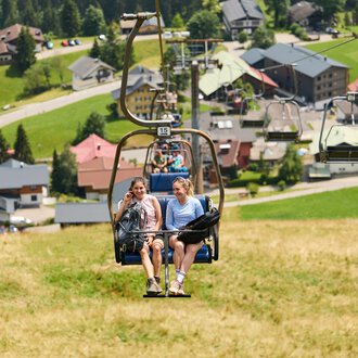 Zwei Frauen sitzen in einer Sesselliftbahn und fahren eine Bergwiese hinauf. Im Hintergrund sind grüne Hügel und ein kleines Dorf mit Häusern zu sehen. | © Kleinwalsertal Tourismus | Oliver Farys