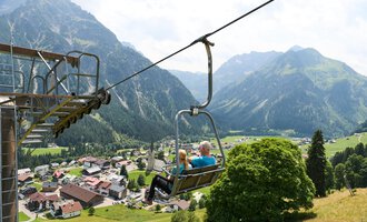 Eine Gondelbahn fährt über ein malerisches Tal mit Bergen im Hintergrund. Im Vordergrund ist ein uriger Ort mit roten Dächern und grünen Wiesen zu sehen. | © Kleinwalsertal Tourismus | Oliver Farys