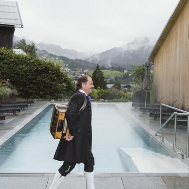 A man in traditional attire is carrying a box and walking along a pool. In the background, mountains and fog can be seen.