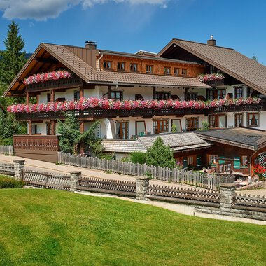 A beautiful alpine hotel with colorful flowers on the balcony. Surrounded by green meadows and trees under a blue sky.